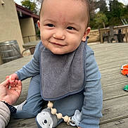 Merrick is registered to the contest to win money with this photo: baby, child, smiling, curly_hair, blue_clothing, bib, pacifier, outdoor, wooden_table, hand_holding, cute, infant, toy, person, skin, face, happy, sitting, daylight, nature