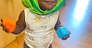 Ever is registered to the contest to win money with this photo: toddler, child, watermelon_hat, toy_block, orange_block, blue_block, indoor, wooden_floor, smiling, playful, sleeveless_shirt, shorts, sandals, person, cute, happy, face, headwear, young_child, home