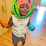 toddler, child, watermelon_hat, toy_block, orange_block, blue_block, indoor, wooden_floor, smiling, playful, sleeveless_shirt, shorts, sandals, person, cute, happy, face, headwear, young_child, home