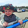 toddler, child, shopping_cart, hat, dinosaur_pattern, smile, outdoor, parking_lot, car, blue_handle, bib, long_sleeve, happy, daylight, clothing, hand, face, trees, sky, seat