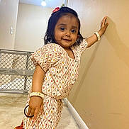 Viha is registered to the contest to win money with this photo: toddler, child, girl, traditional_clothing, bracelet, flower_hair_accessory, carpet, wall, baby_gate, indoor, smiling, standing, hand_on_wall, person, accessory, cute, young_child, home, portrait, fashion