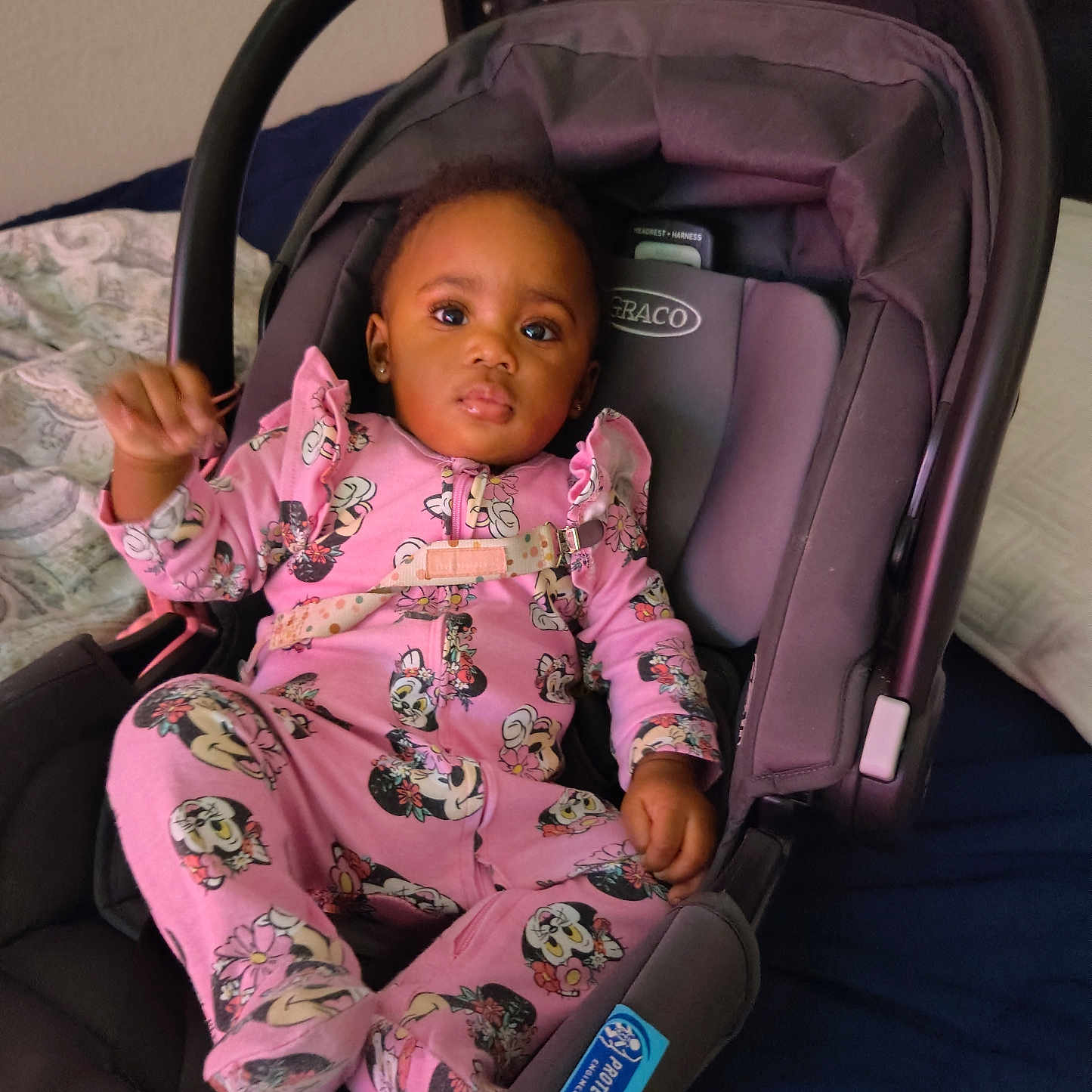 Nya'Lease is registered to the contest to win money with this photo: baby, child, infant, car_seat, graco, pink_clothing, onesie, minnie_mouse, face, earrings, bed, blanket, patterned_sheet, indoor, person, seat_belt, head, expression, cute, portrait