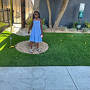 Samiyah is registered to the contest to win money with this photo: child, smiling, outdoor, blue_dress, standing, portrait, artificial_grass, tree, fence, playground, pebbles, sandals, bracelet, necklace, toy, pet_waste_station, garden, sunlight, sidewalk, happy