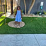 child, smiling, outdoor, blue_dress, standing, portrait, artificial_grass, tree, fence, playground, pebbles, sandals, bracelet, necklace, toy, pet_waste_station, garden, sunlight, sidewalk, happy