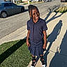 backpack, braids, cars, child, daylight, fashion, grass, happy, houses, outdoor, portrait, school_uniform, shadow, shoes, sidewalk, smiling, standing, street, suburban, sunny