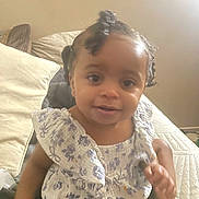 Ryyah is registered to the contest to win money with this photo: baby, child, infant, portrait, smiling, sitting, bed, bedroom, pillow, blanket, dress, floral_pattern, braids, hair_bows, finger_pointing, indoor, cozy, warm_lighting, person, photograph