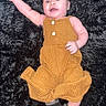 Xylin is registered to the contest to win money with this photo: baby, child, infant, smiling, happy, lying_down, portrait, face, blue_eyes, mustard_outfit, overalls, dress, buttons, black_rug, fluffy_texture, shoes, adult_legs, indoor, wooden_floor, photography