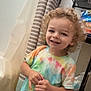 child, toddler, curly_hair, smiling, tie_dye_shirt, colorful_clothing, indoor, curtains, shelf, household_items, pants, patterned_pants, standing, happy, person, young_child, casual_clothing, playful, cute, portrait