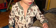 Maxton is registered to the contest to win money with this photo: toddler, child, pajamas, curly_hair, fruit, plate, blueberries, pineapple, strawberry, indoor, couch, wooden_floor, toy, blanket, table, face, person, home, clothing, food