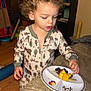 toddler, child, pajamas, curly_hair, fruit, plate, blueberries, pineapple, strawberry, indoor, couch, wooden_floor, toy, blanket, table, face, person, home, clothing, food