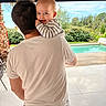 baby, adult, person, smile, outdoor, pool, greenery, trees, sky, clothing, back_view, holding, happy, daylight, casual, nature, patio, relaxation, family, portrait