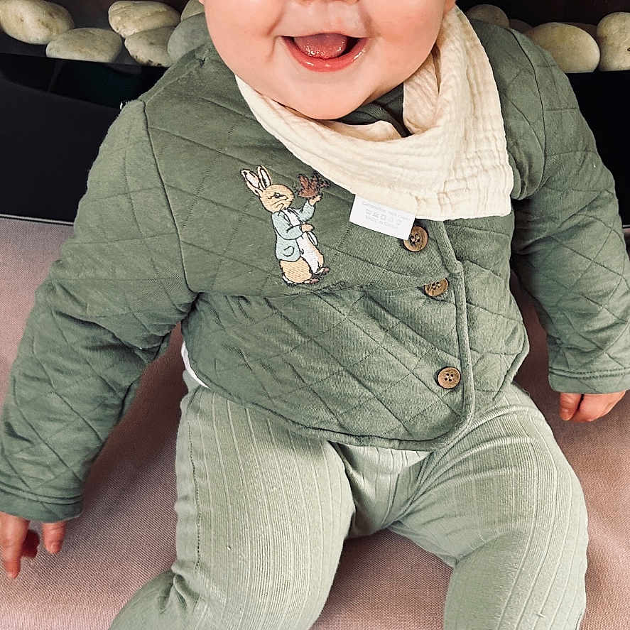 Keegi is registered to the contest to win money with this photo: baby, bunny_patch, buttoned_jacket, child, couch, cozy, cute, eyes, face, green_clothing, happy, head, indoor, infant, portrait, seated, smiling, socks, soft_furniture, textile