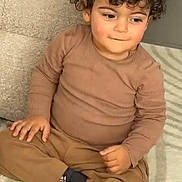 Amir participe au concours pour gagner de l'argent avec cette photo : child, toddler, curly_hair, brown_clothing, socks, carpet, indoor, sitting, cross_legged, cute, face, person, young, floor, portrait, expression, cozy, casual_wear, home, thoughtful