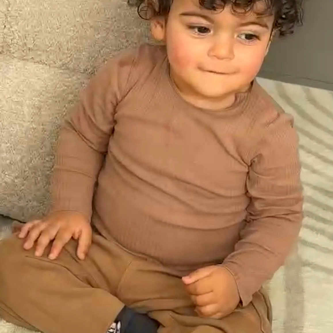 Amir participe au concours pour gagner de l'argent avec cette photo : child, toddler, curly_hair, brown_clothing, socks, carpet, indoor, sitting, cross_legged, cute, face, person, young, floor, portrait, expression, cozy, casual_wear, home, thoughtful
