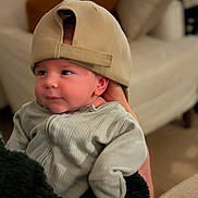 Frederick is registered to the contest to win money with this photo: baby, infant, cap, hat, clothing, person, indoor, portrait, cute, child, holding, cozy, soft_focus, furniture, adult_hand, looking_away, seated, warm_lighting, smiling, casual