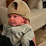 baby, infant, cap, hat, clothing, person, indoor, portrait, cute, child, holding, cozy, soft_focus, furniture, adult_hand, looking_away, seated, warm_lighting, smiling, casual