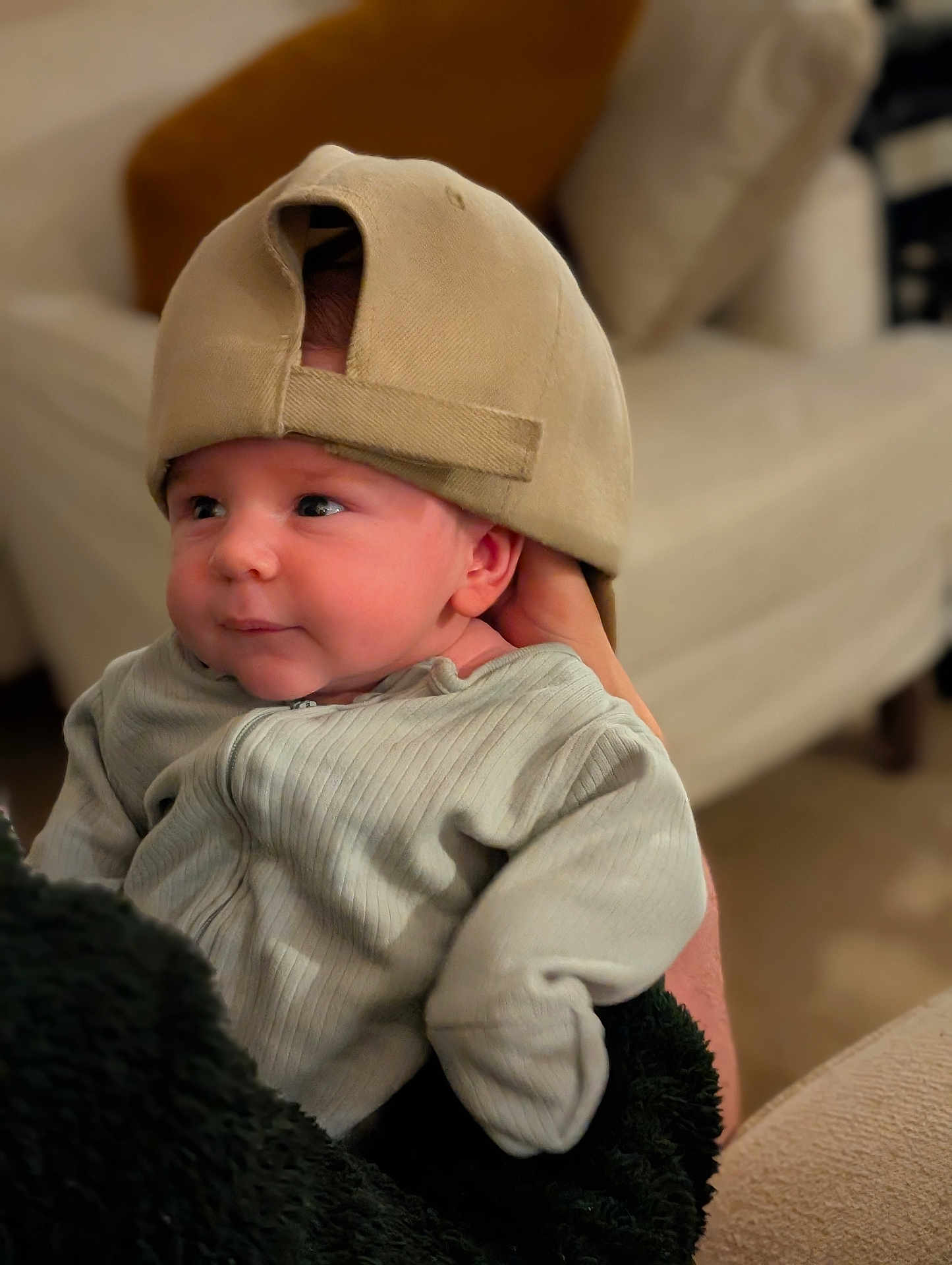 Frederick is registered to the contest to win money with this photo: baby, infant, cap, hat, clothing, person, indoor, portrait, cute, child, holding, cozy, soft_focus, furniture, adult_hand, looking_away, seated, warm_lighting, smiling, casual