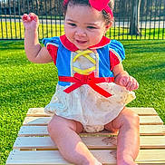 Storm is registered to the contest to win money with this photo: child, baby, toddler, smiling, red_bow, headband, dress, snow_white_costume, sitting, wooden_crate, grass, outdoor, park, sunlight, fence, portrait, feet, legs, hand_raised, cute