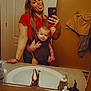 Braxton is registered to the contest to win money with this photo: woman, baby, mirror, bathroom, selfie, phone, sink, toiletries, towel, red_shirt, baby_carrier, face, tongue_out, tattoo, mirror_reflection, digital_filter, devil_horns, playful, smile, casual