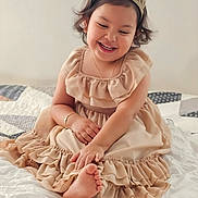Camila is registered to the contest to win money with this photo: child, girl, smiling, crown, dress, beige, bed, blanket, happy, cute, sitting, foot, playful, indoors, portrait, curly_hair, jewelry, bracelet, cozy, soft_lighting