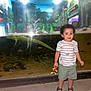 toddler, child, aquarium, fish, alligator, toy, curly_hair, shorts, striped_shirt, sandals, glass_tank, underwater, jungle_mural, bright_lights, reflection, indoor, smiling, standing, playful, nature_theme