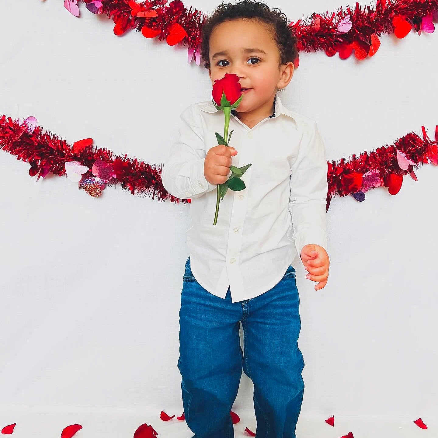 Neiko is registered to the contest to win money with this photo: blue_jeans, celebration, child, cute, decorations, flower, happy, holiday, indoor, person, petals, portrait, red_garland, red_rose, rose, smiling, standing, toddler, valentines_day, white_shirt
