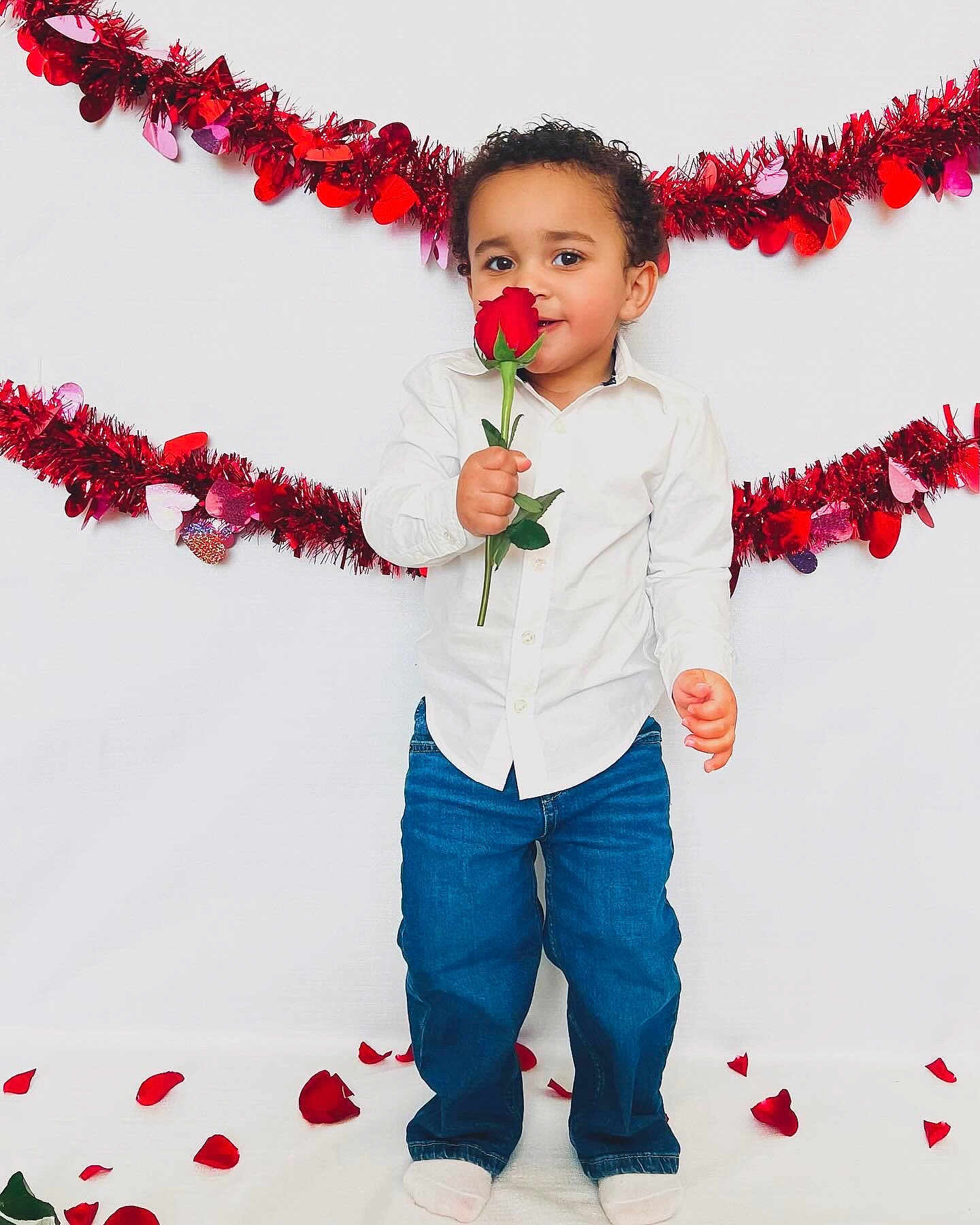 Neiko is registered to the contest to win money with this photo: toddler, child, rose, red_rose, valentines_day, flower, petals, white_shirt, blue_jeans, smiling, standing, indoor, decorations, red_garland, cute, portrait, holiday, celebration, happy, person