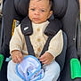baby, child, car_seat, blanket, sweater, footed_pants, seatbelt, straps, outdoor, sidewalk, car_tire, green_handle, knitwear, infant, young_child, serious_face, person, clothing, sitting, baby_accessory