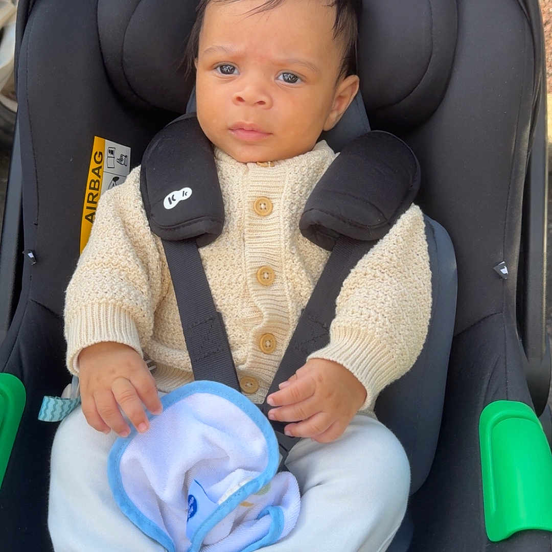 Léo a rejoint le concours — aidez-le/la à gagner de superbes lots ! baby, baby_accessory, blanket, car_seat, car_tire, child, clothing, footed_pants, green_handle, infant, knitwear, outdoor, person, seatbelt, serious_face, sidewalk, sitting, straps, sweater, young_child