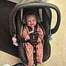 baby, infant, car_seat, smiling, child, onesie, floral_pattern, indoor, flooring, carpet, tile, footwear, black_car_seat, safety_harness, happy, person, cute, seated, head_support, playful