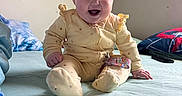 Hazel joined the competition — help win amazing prizes! baby, bed, blanket, cozy, cute, face, foot, hair, hand, happy, indoor, infant, person, pillow, room, sitting, smile, text, wall_banner, yellow_onesie