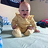 Hazel joined the competition — help win amazing prizes! baby, infant, yellow_onesie, bed, pillow, smile, happy, indoor, face, person, wall_banner, text, room, hand, foot, blanket, hair, sitting, cute, cozy