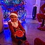 baby, child, christmas_tree, christmas_lights, santa_hat, santa_outfit, toy, spiderman, ball, floor, tile_floor, festive_clothing, holiday, indoor, person, smile, decoration, red_clothing, blue_lights, happy