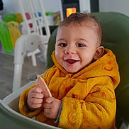 Noham a rejoint le concours — aidez-le/la à gagner de superbes lots ! baby, child, high_chair, yellow_jacket, smiling, happy, indoor, cozy, food, snack, warm, furniture, toy, fireplace, face, hands, portrait, cute, infant, clothing