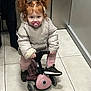 child, cute, hallway, indoor, jacket, pacifier, person, pigtails, pink_pants, play, red_hair, ride_on, small_vehicle, sneakers, standing, tile_floor, toddler, toy, warm_clothing, young_child