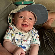 Hunter is registered to the contest to win money with this photo: adult_arm, baby, baseball_cap, casual, child, clothing, cute, drool, happy, hat, home, indoor, infant, living_room, person, portrait, relaxing, sitting, smiling, sofa