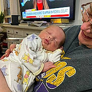 Samuel is registered to the contest to win money with this photo: baby, sleeping, pajamas, winnie_the_pooh, person, smiling, television, news, indoor, living_room, couch, sweatshirt, glasses, holding, child, cute, resting, face, human, home