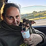 person, dog, car_interior, window, cup, holding, sunlight, casual_clothing, closeup, daylight, road, trees, outdoor_view, pet, tongue_out, face, hand, sitting, animal, transportation