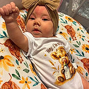 Hadley is registered to the contest to win money with this photo: baby, infant, headband, onesie, cushion, floral_pattern, cartoon_cow, raised_fist, portrait, cute, child, animal_print, indoor, clothing, baby_girl, expression, face, newborn, soft_toys, colorful