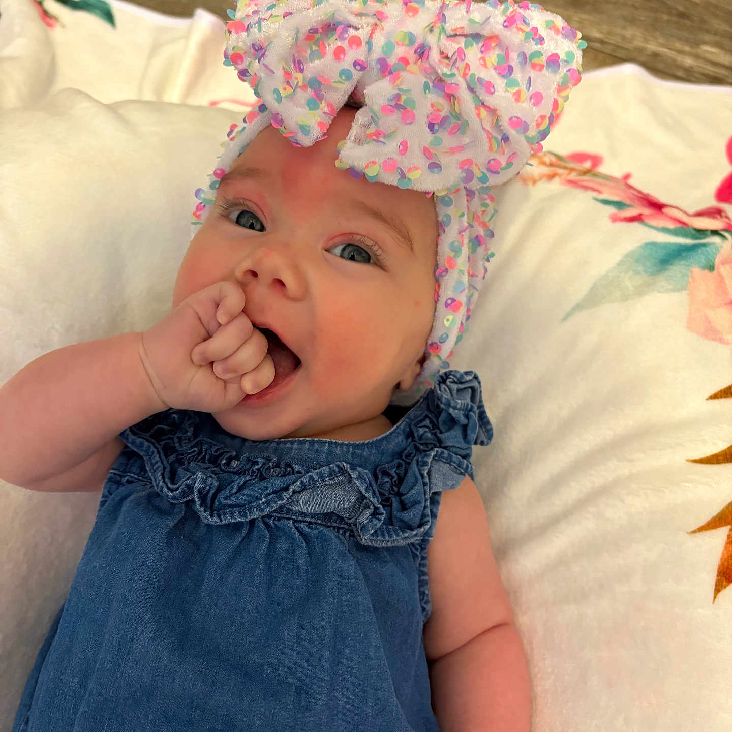 Hadley is registered to the contest to win money with this photo: baby, blue_dress, bow, child, closeup, cushion, cute, floral_pattern, happy, headband, headwear, indoors, infant, person, portrait, relaxing, skin, smiling, soft_texture, thumb_sucking