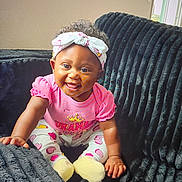 Italy Rose is registered to the contest to win money with this photo: baby, child, smiling, pink_clothing, headband, couch, indoor, happy, sitting, curly_hair, yellow_socks, heart_pattern, infant, cute, portrait, furniture, person, cozy, playful, room