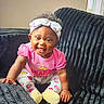 baby, child, smiling, pink_clothing, headband, couch, indoor, happy, sitting, curly_hair, yellow_socks, heart_pattern, infant, cute, portrait, furniture, person, cozy, playful, room