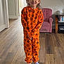 child, smiling, pajamas, orange_clothing, pumpkin_pattern, living_room, wooden_floor, pink_armchair, socks, happy, indoor, person, home, television, furniture, flooring, cozy, cute, portrait, casual