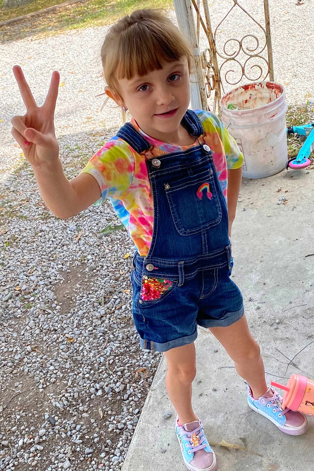 Lillian is registered to the contest to win money with this photo: child, girl, peace_sign, tie_dye, denim_overalls, shorts, sneakers, outdoor, porch, gravel, concrete, cup, scooter, smile, playful, casual_clothing, summer, young, happy, posing