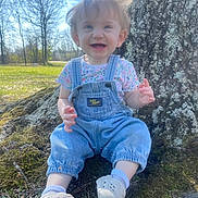 Nova is registered to the contest to win money with this photo: baby, blue_sky, casual_clothing, child, daytime, denim_overalls, face, grass, happy, moss, nature, outdoor, park, person, portrait, shoes, smiling, sunlight, toddler, tree