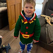 Tyler is registered to the contest to win money with this photo: child, smiling, indoor, green_sweater, red_buttons, yellow_accents, blue_pants, sneakers, wooden_floor, toys, door, couch, blanket, happy, person, home, casual_clothing, cute, young, standing