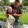 baby, man, smiling, outdoor, backyard, grass, table, blanket, hat, socks, glasses, fence, plants, shirt, holding, happy, person, nature, casual, family