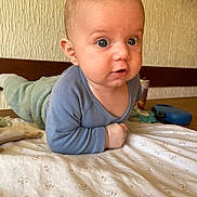 Téo a rejoint le concours — aidez-le/la à gagner de superbes lots ! baby, child, infant, blue_clothing, green_pants, blanket, patterned_blanket, lying_down, curious_expression, indoors, toys, wooden_surface, face, wide_eyes, propped_up, elbows, head, skin, person, young_child