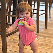Amira joined the competition — help win amazing prizes! toddler, child, baby, pink_onesie, snack_pouch, wooden_chair, wooden_floor, indoor, standing, holding, cute, chubby_legs, polka_dot, furniture, flooring, young_child, candid, portrait, playful, expression