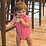toddler, child, baby, pink_onesie, snack_pouch, wooden_chair, wooden_floor, indoor, standing, holding, cute, chubby_legs, polka_dot, furniture, flooring, young_child, candid, portrait, playful, expression